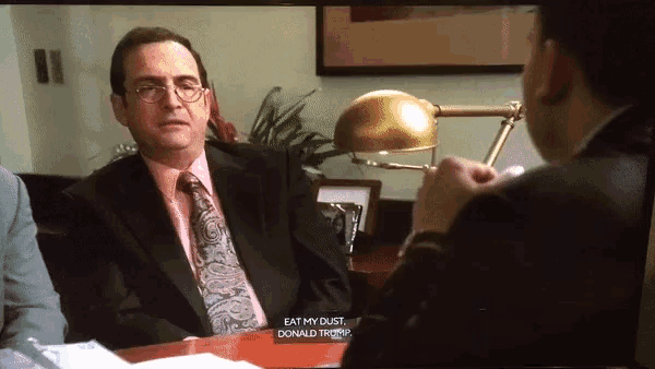 a man in a suit and tie is sitting at a table with a sign that says eat my dust