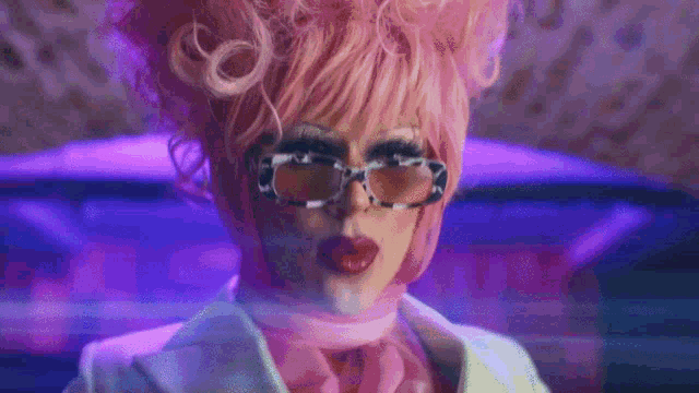 a drag queen wearing a pink wig and sunglasses is sitting on a bed .