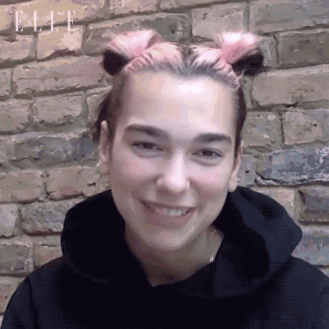 a woman with pink hair is wearing a black hoodie and smiling in front of a brick wall