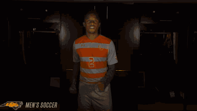 a man in an orange shirt is standing in a dark room with the words men 's soccer behind him