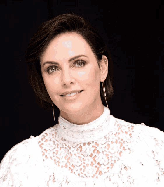 a woman wearing a white lace top and earrings smiles at the camera
