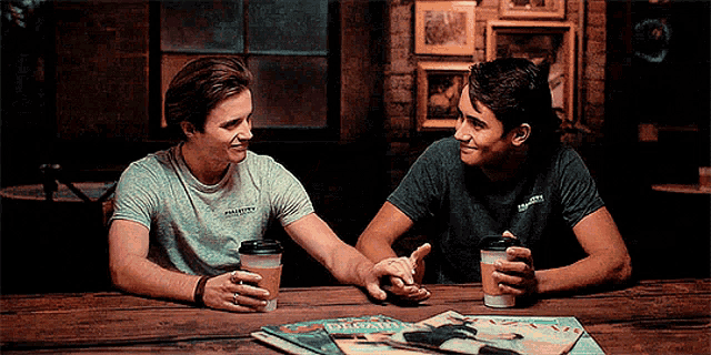 two men are sitting at a table holding hands and one has a shirt that says ' converse ' on it
