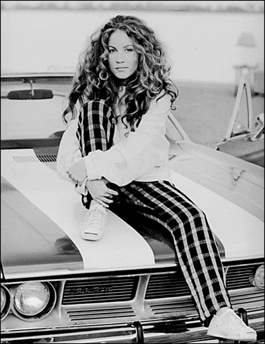 a woman in plaid pants sits on the hood of a muscle car