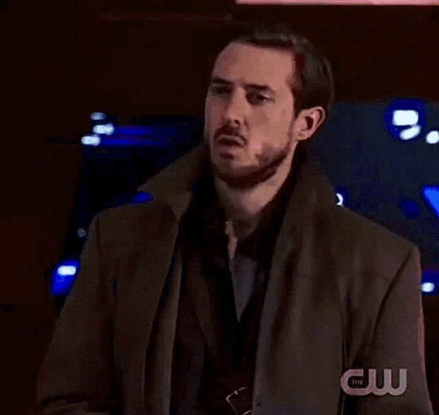 a man in a trench coat is standing in front of a cw screen