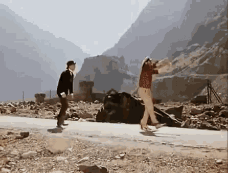 a man and a woman are walking down a road in front of mountains .