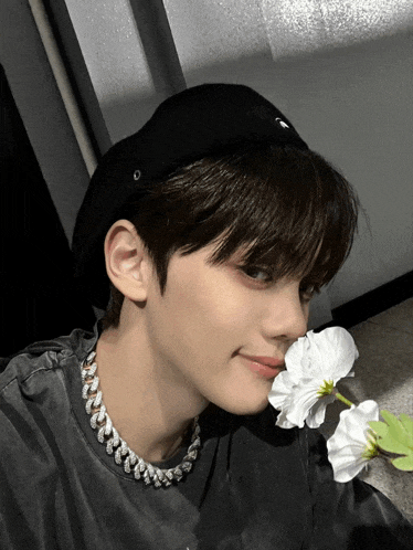 a boy wearing a black hat and a necklace smells a flower