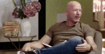a bald man is sitting on a couch with his legs crossed and a vase of flowers in the background .