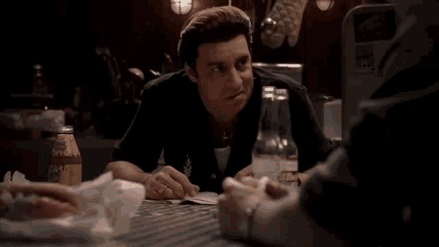 a man sitting at a table talking to another man with a bottle of soda on the table