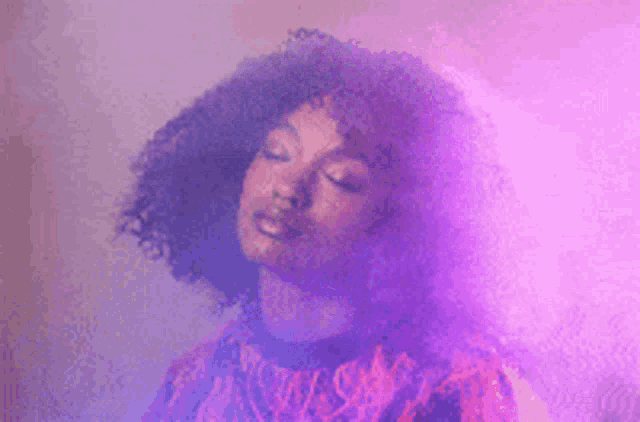 a woman with curly hair is standing in front of a purple smoke .
