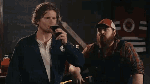 a man is drinking a beer while standing next to another man with a beard .