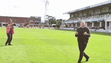 a man in a black jacket is running on a soccer field in front of a stadium