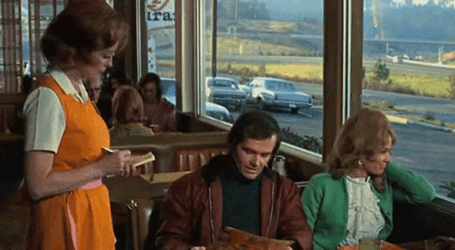 a woman in an orange apron is taking an order from a man and woman in a diner