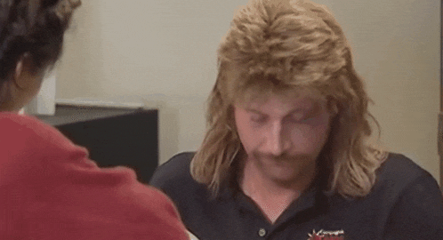 a man with a mullet and mustache is talking to a woman in a red shirt .