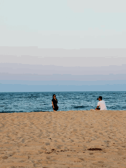 a man sits on the beach while a woman stands on the sand
