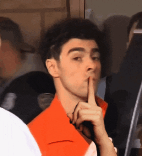 a man in an orange shirt holds his finger to his lips in a gesture of silence