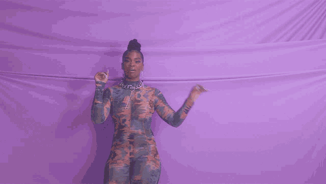 a woman in a camouflage jumpsuit is dancing in front of a purple cloth