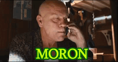 a bald man is talking on a cell phone and the word moron is on the screen behind him