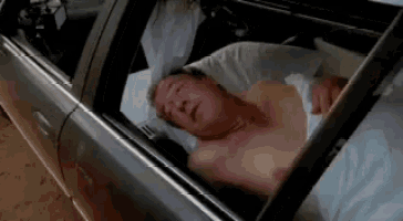 a shirtless man is sleeping in a car with his head out the window .