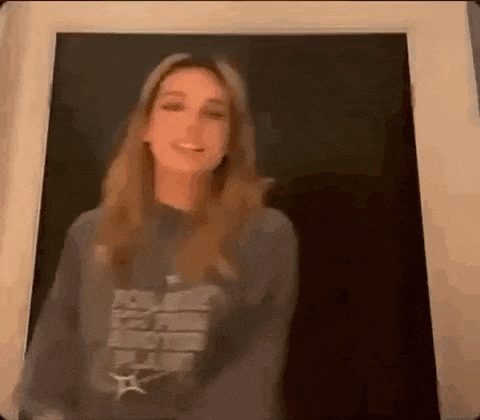 a woman in a gray sweatshirt is standing in front of a black wall and smiling .