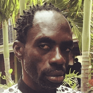 a man with dreadlocks and a beard is standing in front of palm trees and looking at the camera .