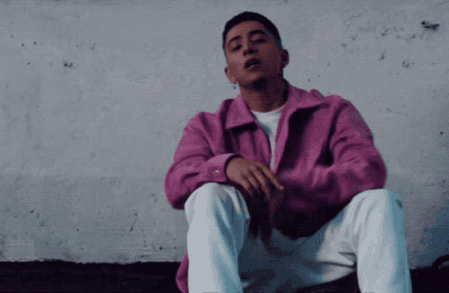 a man in a pink jacket and white pants sits in front of a wall