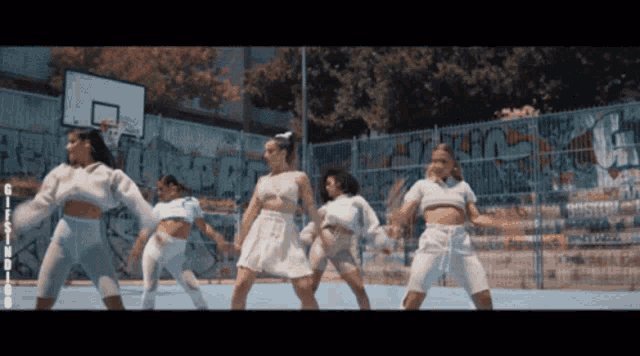 a group of women are dancing in front of a basketball hoop and a fence with graffiti on it