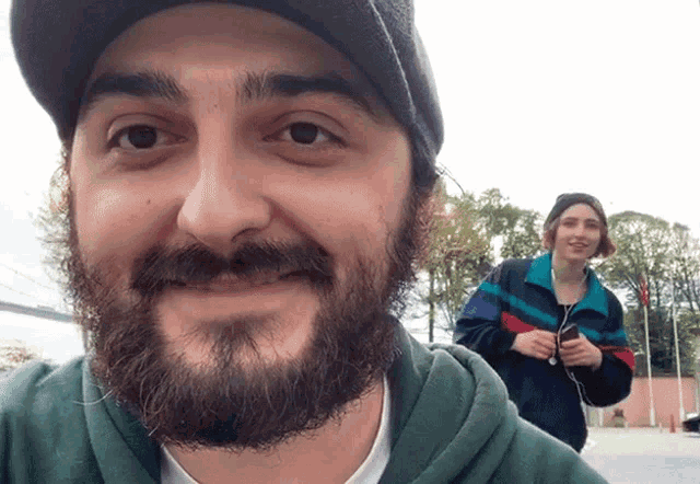a man with a beard is smiling while a woman stands behind him holding a cell phone