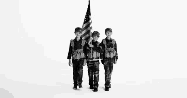 three young boys in military uniforms are walking with an american flag in a black and white photo .