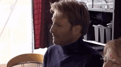 a man wearing a black turtleneck sweater is sitting in front of a shelf with containers on it .