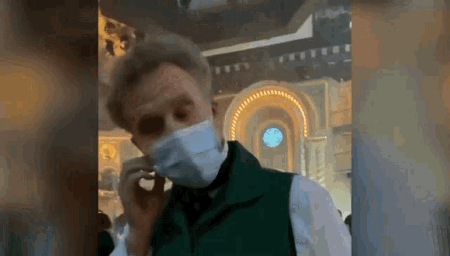 a man wearing a face mask and a green vest is talking on a cell phone .