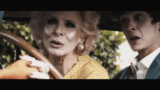 a woman in a yellow polka dot shirt is driving a car next to a man