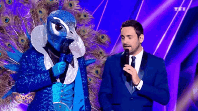 a man in a suit and tie stands next to a peacock masked person holding a microphone