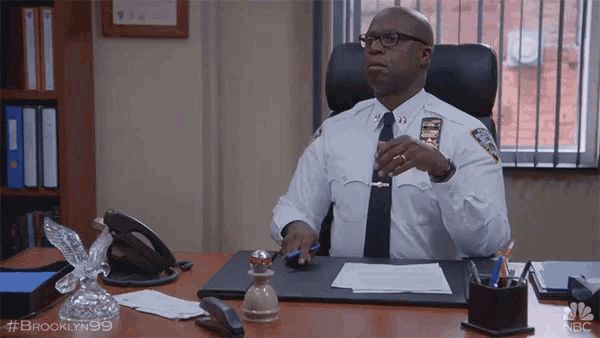 a man in a police uniform sits at a desk with the hashtag #brooklyn99 on the bottom