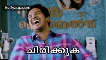 a man in a plaid shirt is laughing in front of a wall that says ' malayalam ' on it