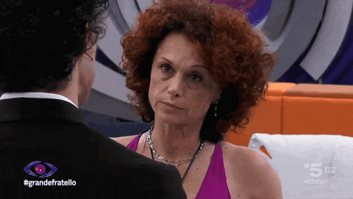 a woman with red hair looks at a man in a tuxedo in front of a sign that says grande fratello