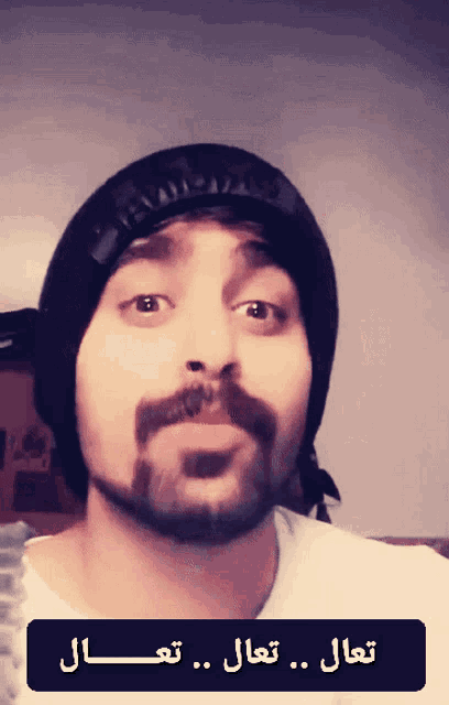 a man with a beard wearing a black beanie and a white shirt with arabic writing on it