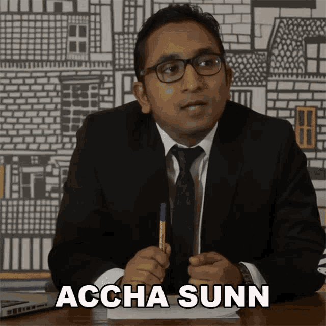 a man in a suit and tie is sitting at a desk with the words accha sunn written on the screen behind him