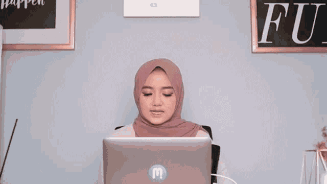 a woman wearing a hijab sits in front of a laptop with the word khawatir written on the wall behind her