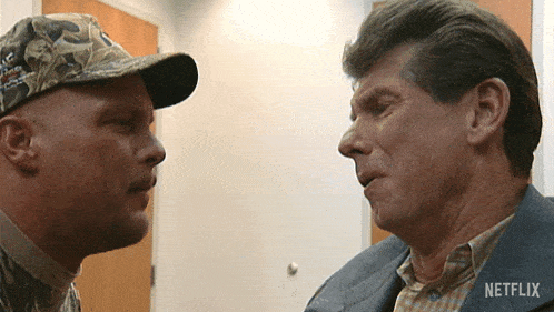 a man in a camo hat talks to another man in a suit with netflix written on his shirt