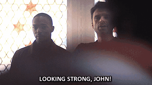two men are standing in front of a window and one of them is saying looking strong john