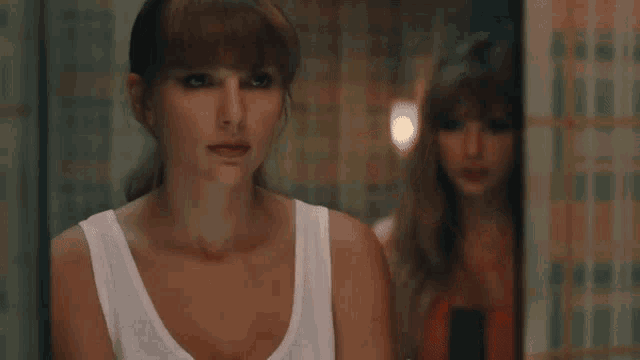 a woman in a white tank top is looking at herself in the mirror .