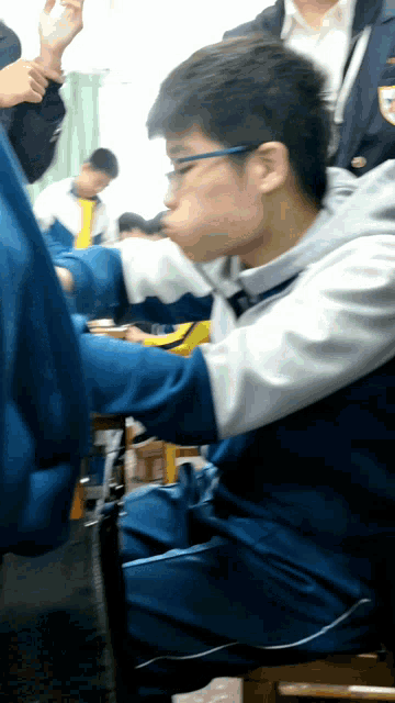 a young man wearing glasses sits in a classroom with other students