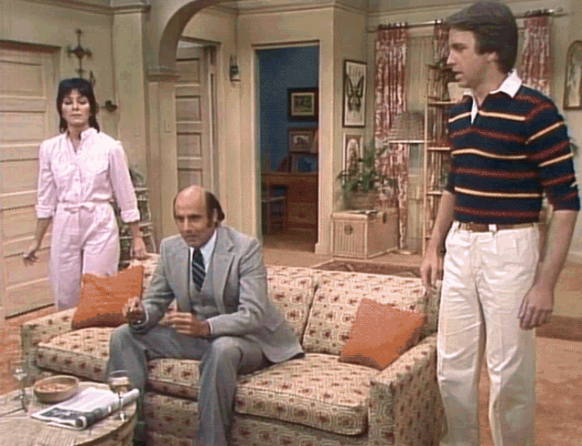 a man in a suit sits on a couch with a woman standing behind him