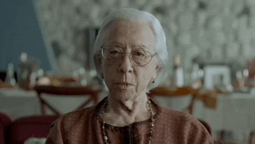 an elderly woman wearing glasses and a necklace is looking at the camera