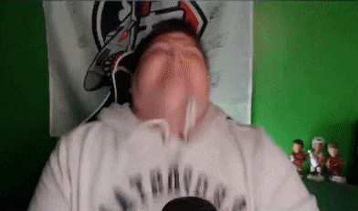 a man is making a funny face in front of a green wall while wearing a patriots sweatshirt .
