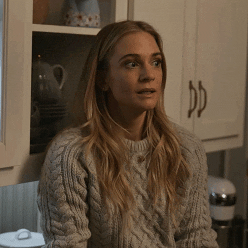 a woman in a grey sweater sits in front of a cabinet
