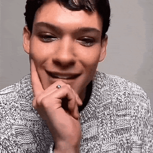 a man wearing a sweater and a ring on his finger is smiling with his hand on his chin .