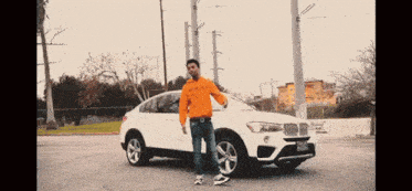 a man in an orange sweatshirt stands in front of a white bmw