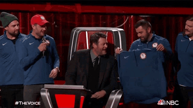 a group of men are standing around a man holding a shirt with the word nbc on it
