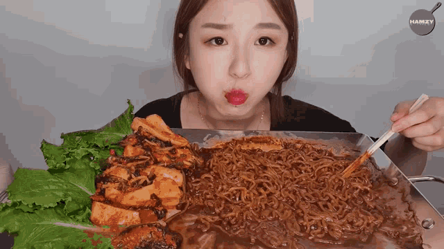 a woman is eating noodles and lettuce with chopsticks from a pan that says hamxy on the bottom